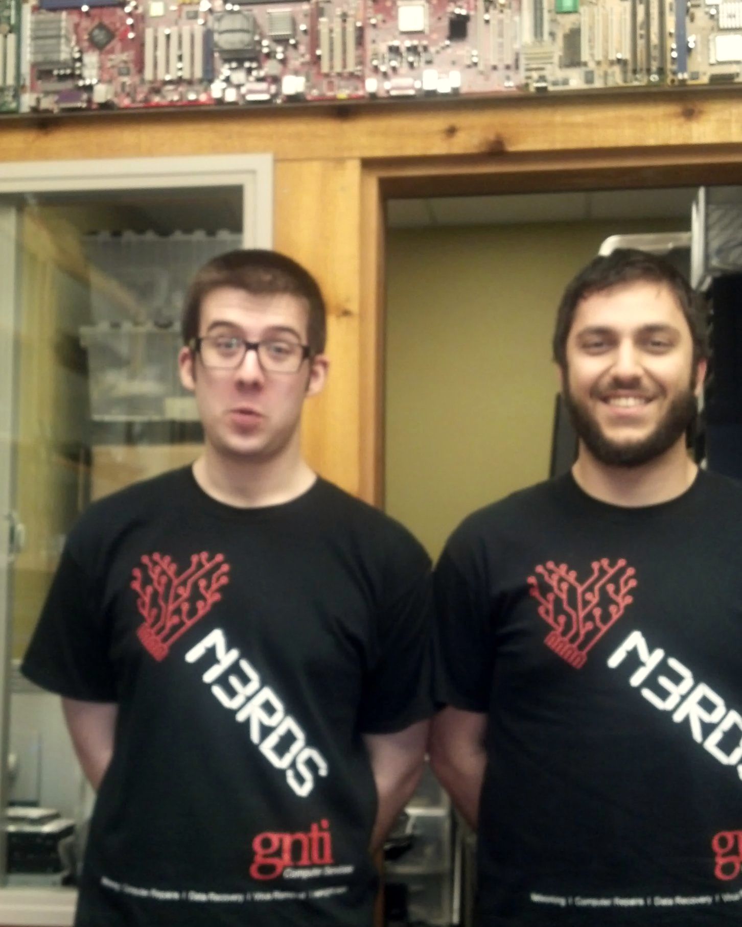 Two GNTI technicians in matching GNTI t-shirts in front of a wall of motherboards at the GNTI shop